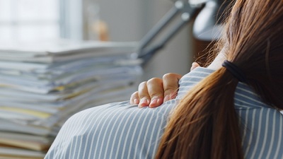 Jeune femme se tenant le haut du dos, devant son bureau rempli de dossiers