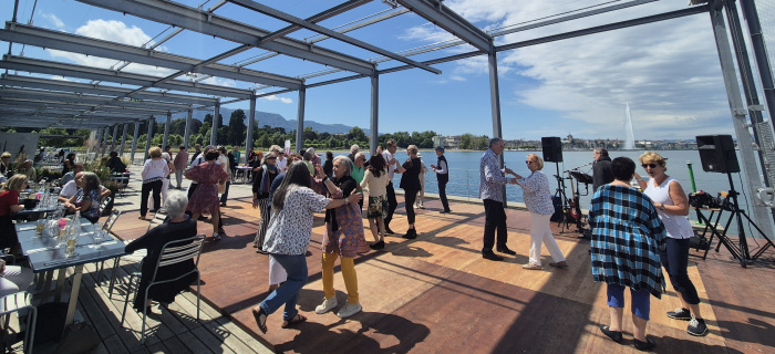 Photographie de seniors dansant sur la terrasse de la Plage des Eaux-Vives, avec le jet d'eau en fond © Hg