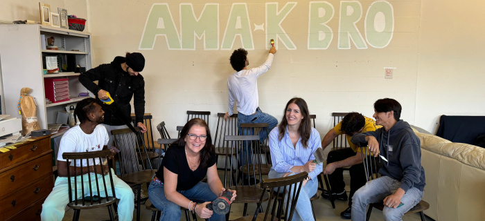Plusieurs personnes sont assises et sourient dans une salle. A l'arrière, un jeune peint sur le mur le logo d'AMA-K BRO log