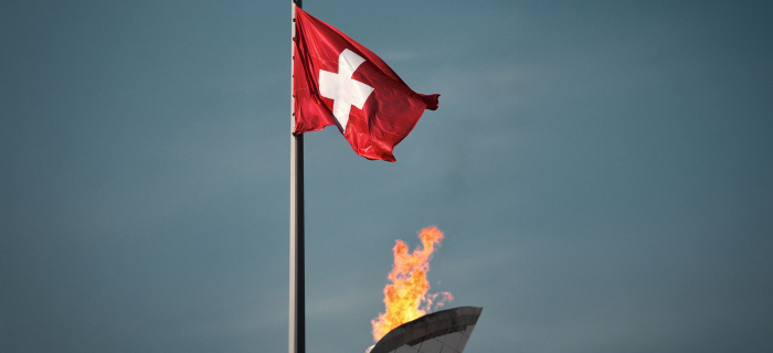 La flamme olympique brûle avec, sur sa gauche, le drapeau suisse flottant au vent