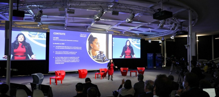 Salle de conférence avec public nombreux. Une femme s'exprime assise sur scène, avec des projections diverses derrière elle.