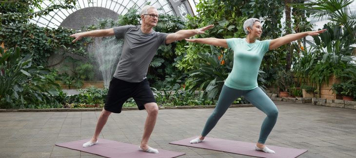 Un homme et une femme aux cheveux gris font de l'exercice sur des tapis de de yoga dans une serre de jardin botanique