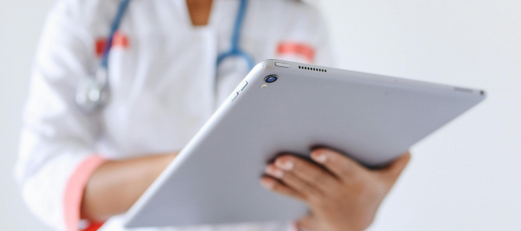 Une femme dont ne voit pas le visage mais on devine qu'elle porte un masque médical est en blouse blanche, avec un stéthoscope autours du coup et tien une tablette qui est en premier plan