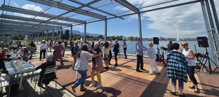 Photographie de seniors dansant sur la terrasse de la Plage des Eaux-Vives, avec le jet d'eau en fond © Hg