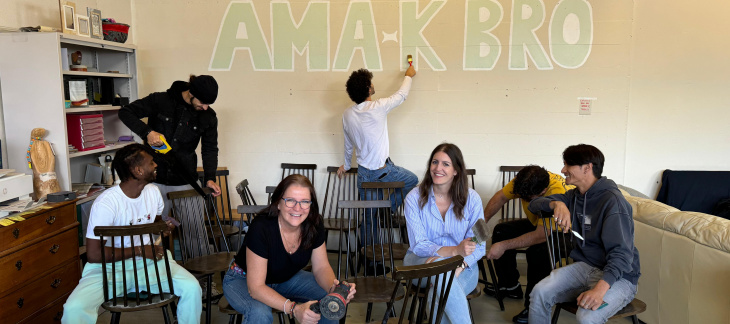 Plusieurs personnes sont assises et sourient dans une salle. A l'arrière, un jeune peint sur le mur le logo d'AMA-K BRO log 