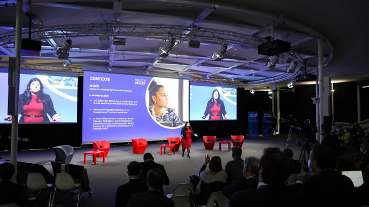 Salle de conférence avec public nombreux. Une femme s'exprime assise sur scène, avec des projections diverses derrière elle.