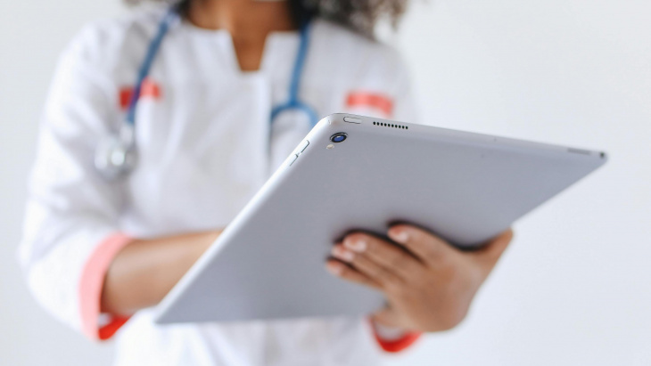 Une femme dont ne voit pas le visage mais on devine qu'elle porte un masque médical est en blouse blanche, avec un stéthoscope autours du coup et tien une tablette qui est en premier plan 