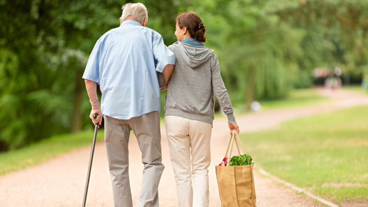 Une femme porte un sac de courses et accompagne un homme plus âgé avec une canne