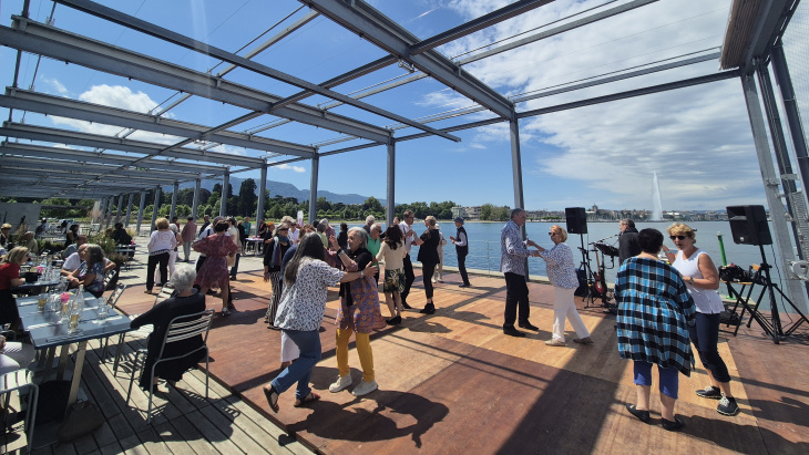 Photographie de seniors dansant sur la terrasse de la Plage des Eaux-Vives, avec le jet d'eau en fond © Hg