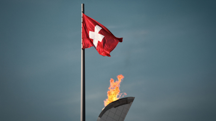 La flamme olympique brûle avec, sur sa gauche, le drapeau suisse flottant au vent